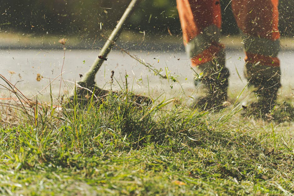 Close-up of a gardener's feet and legs in orange gardening overalls, using a grass rake to clear overgrown grass and weeds from a garden bed area near a pathway. The rake's metal tines are lifting grass clippings and small plant debris, creating a flurry of soil and foliage particles in the air. Surrounding the work area are lush, green garden beds bordered by the lawn, with a mix of wild grasses, small bushes, and clippings scattered across the freshly disturbed soil. Soft natural light illuminates the scene, highlighting the textures of the cut grass, damp soil, and the gardener's protective footwear. This image exemplifies professional garden maintenance and landscaping work carried out by Gardeners Finchley on Ballards Lane, demonstrating attentive hedge trimming and lawn care in a residential garden setting.