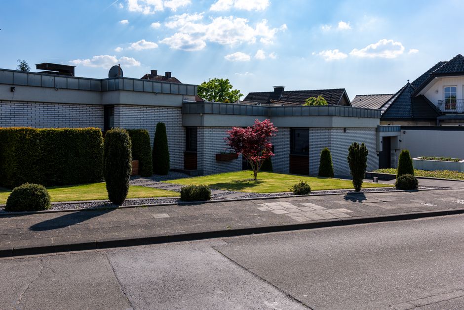 A modern residential building with a flat roof and white brick exterior set behind a landscaped front yard. The garden features a well-maintained lawn edged with neatly trimmed box hedges and dark green conical shrubs. A small, vibrant red-leafed Japanese maple tree adds color to the scene, surrounded by a pathway of dark paving stones. The garden layout is simple yet structured, with distinct sections separated by borders and gravel, showcasing careful garden maintenance and landscaping work. The bright daylight and partly cloudy sky illuminate the scene, emphasizing the textures of the grass, leaves, and paving materials. Gardeners Finchley likely performs lawn care, hedge trimming, and general outdoor maintenance to keep this tidy and aesthetically pleasing outdoor space.
