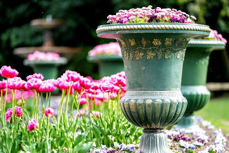 A well-maintained garden features large decorative ceramic pots with ornate floral patterns, filled with blooming pink and purple flowers. Surrounding the pots, there are lush green grass and a variety of vibrant flowers, including poppies with delicate petals and tall, slender stems. The garden layout shows a pathway or flower bed bordered by small purple and white flowers, with a backdrop of mature trees and leafy greenery that provide shade and structure. The soil appears moist and freshly tended, indicating recent garden maintenance by Gardeners Finchley. The scene is illuminated by soft, natural daylight, highlighting the textures of the leaves, flowers, and ceramic pots, reflecting careful landscaping work aimed at enhancing aesthetic appeal.